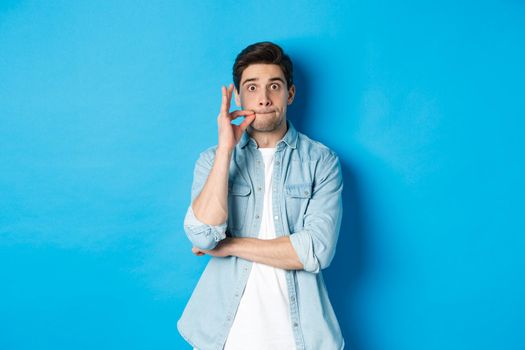 Adult man zipping mouth, promise keep secret, making a seal on lips and standing over blue background.