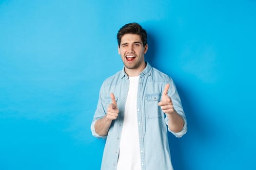 Confident adult man winking, pointing finger at you and winking, congratulating or praising person, standing against blue background.