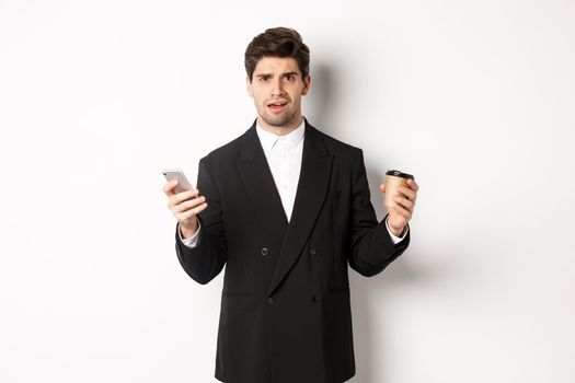 Image of confused businessman standing in a suit, holding cup of coffee and mobile phone, cant understand something, standing against white background.