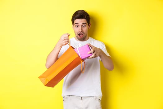 Concept of holidays and celebration. Young man looking surprised as take out gift from shopping bag, standing over yellow background.