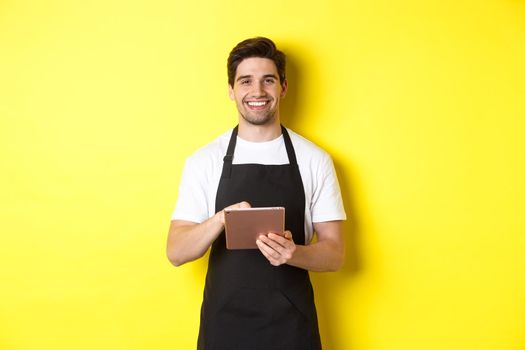 Handsome waiter taking orders, holding digital tablet and smiling, wearing black apron uniform, standing over yellow background.