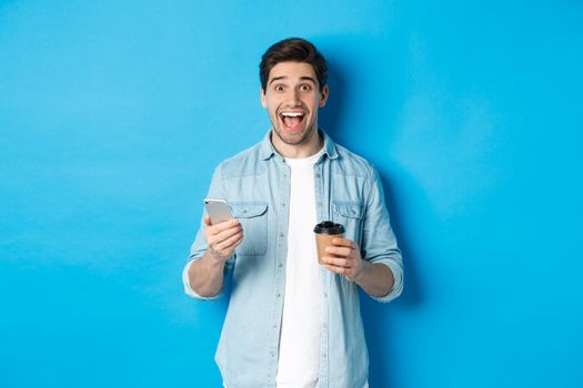 Happy young man drinking coffee and using mobile phone, looking excited, standing against blue background.