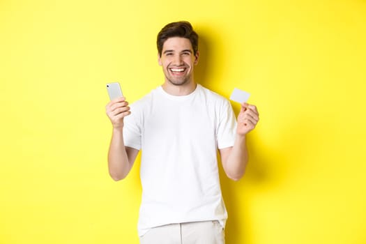 Happy young man shopping online in smartphone, holding credit card and smiling, standing over yellow background.