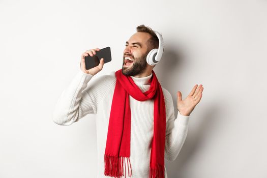 Winter holidays and technology concept. Man playing karaoke app on mobile phone, singing in smartphone, wearing headphones, standing over white background.