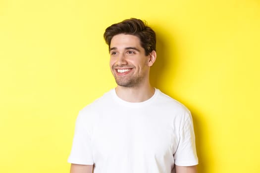 Close-up of attractive bearded man in white t-shirt smiling, looking left at copy space, standing against yellow background.