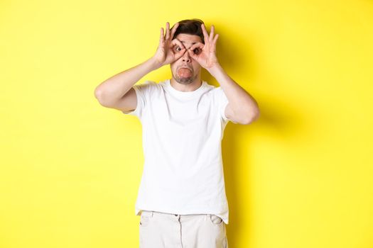 Young man making funny faces and showing tongue, fool around, standing in white t-shirt against yellow background. Copy space