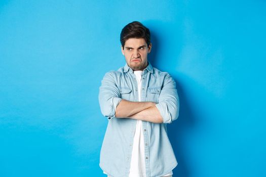 Angry guy cross arms on chest and looking away with insulted expression, standing offended against blue background.
