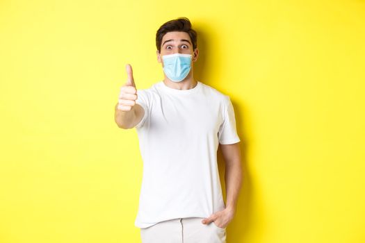 Concept of coronavirus, pandemic and social distancing. Impressed guy in medical mask showing thumb up in approval, like something awesome, yellow background.