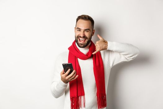 Happy bearded guy holding smartphone, showing phone sign, asking to call him, standing in christmas sweater and scarf, white background.