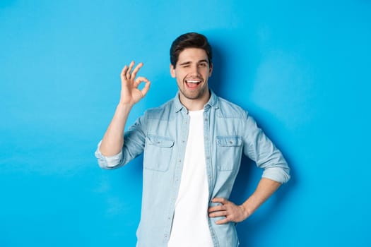 Confident adult man winking and showing okay sign, approve something good, standing against blue background.