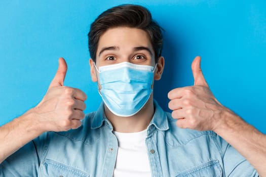 Concept of covid-19, pandemic and quarantine. Close-up of cheerful young man in medical mask smiling, showing thumbs up in approval, like and agree, standing over blue background.