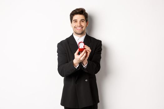 Image of handsome boyfriend in black suit making a proposal, asking to marry him and showing wedding ring, standing over white background.