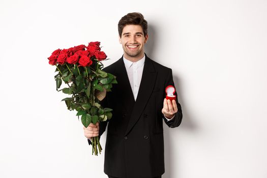 Handsome smiling man in black suit, holding roses and engagement ring, making a proposal to marry him, standing against white background.