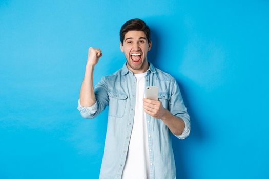 Concept of online shopping, applications and technology. Excited man shouting yes and making fist pump gesture after winning on smartphone, standing over blue background.