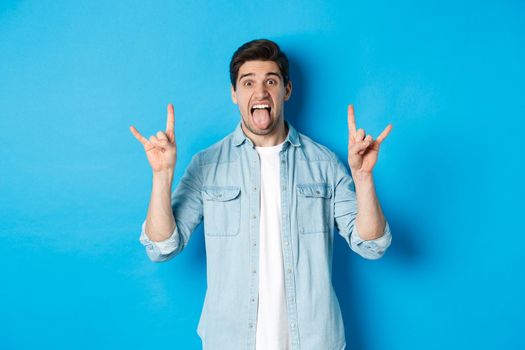 Cool guy having fun, enjoying concert, showing rock sign and tongue, standing over blue background.