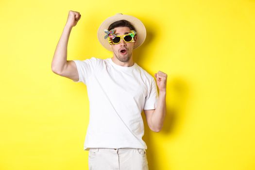 Concept of tourism and lifestyle. Happy guy tourist enjoying trip, rooting for you, fist pump and triumphing, going on journey in summer hat and sunglasses, yellow background.