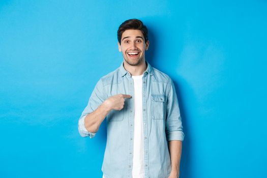 Happy and surprised man pointing at himself, smiling pleased, standing against blue background.