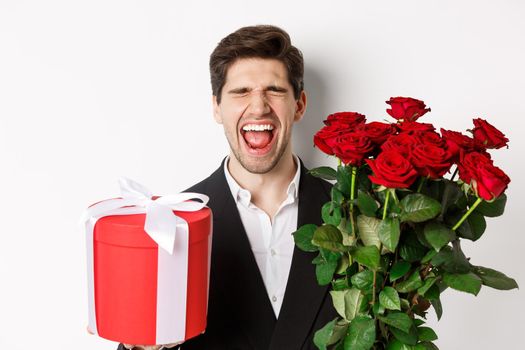 Image of sad guy in suit, got rejected and crying, holding bouquet of roses and present, standing miserable against white background.