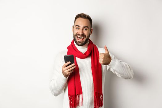 Satisfied man seeing something good online, showing thumbs-up while using mobile phone, standing over white background.