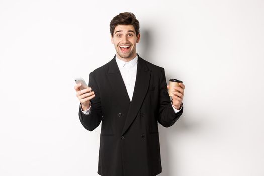 Portrait of happy good-looking man in suit, holding cup of coffee and smartphone, achieve app goal, standing over white background.