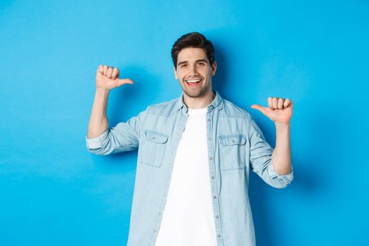 Portrait of confident man pointing at himself, being a professional, bragging while standing against blue background.