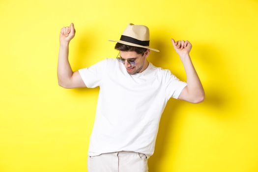Tourism, travelling and holidays concept. Man tourist enjoying vacation, dancing in straw hat and sunglasses, posing against yellow background.
