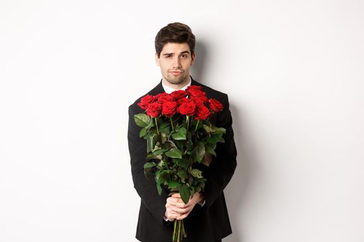 Image of handsome boyfriend in black suit, holding bouquet of red roses and smiling, being on a date, standing over white background.