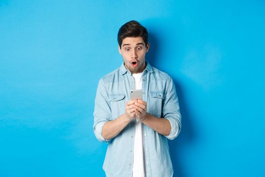 Man looking amazed while checking promo on smartphone, looking surprised at phone, standing against blue background.