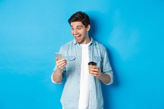 Man looking surprised at smartphone, drinking coffee, standing over blue background amazed.