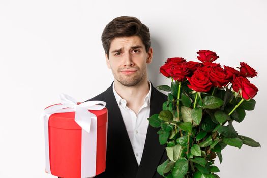 Close-up of sad man in suit, holding bouquet of red roses and a gift, standing upset against white background.