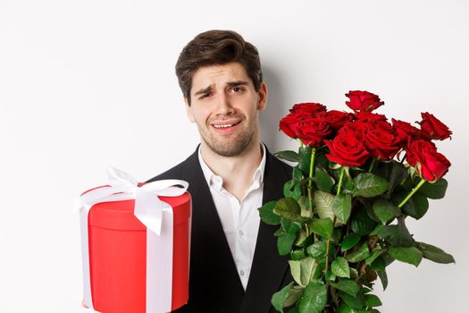 Close-up of skeptical man in suit, holding bouquet of red roses and a gift, standing reluctant against white background.