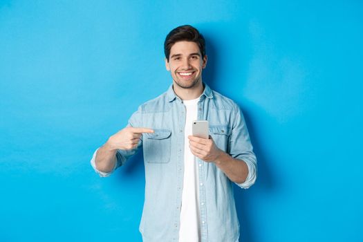 Concept of online shopping, applications and technology. Handsome man recommending app on smartphone, pointing at phone and smiling satisfied, standing over blue background.