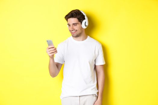 Handsome modern guy choosing playlist on smartphone, wearing headphones, standing over yellow background.