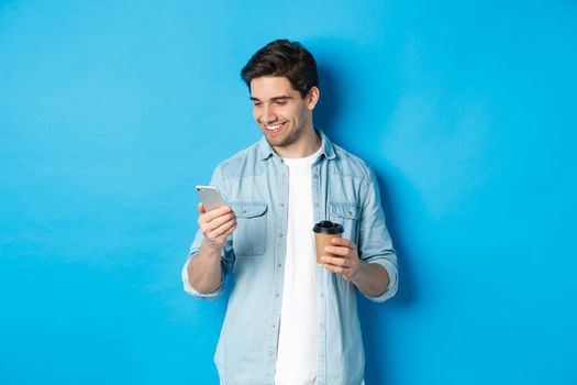 Handsome modern guy reading message on cell phone and drinking coffee, standing against blue background.