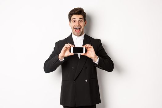 Image of cheerful, handsome man in black suit, showing smarthone screen and looking amazed, standing against white background.