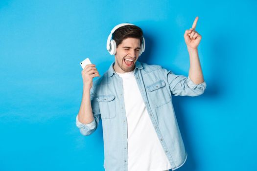 Happy handsome man dancing, listening music on smartphone in headphones, standing over blue background.