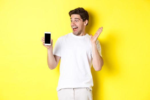 Image of amazed guy looking at mobile phone screen with surprised face, standing excited against yellow background.