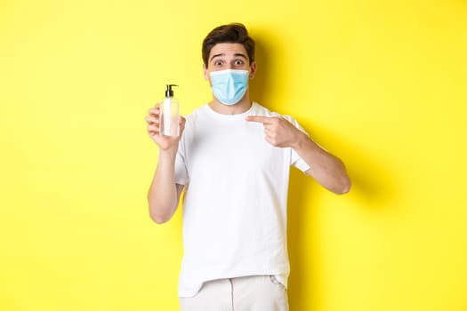 Concept of covid-19, quarantine and lifestyle. Excited guy in medical mask showing good hand sanitizer, pointing finger at antiseptic, standing over yellow background.