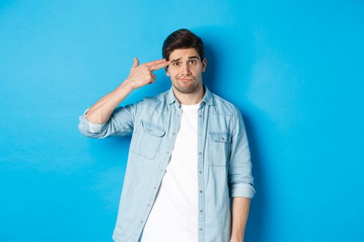 Tired and annoyed adult man making finger gun gesture near head, shooting himself from boredom, standing against blue background.