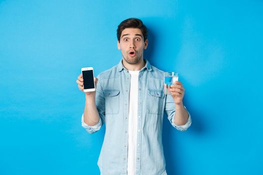 Man looking surprised, showing smartphone screen and glass of water, standing over blue background.