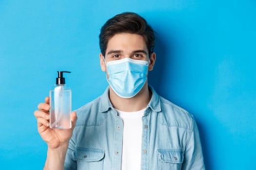Concept of coronavirus, pandemic and preventive measures. Close-up of young man in medical mask advice to use hand sanitizer, showing antiseptic bottle, standing over blue background.