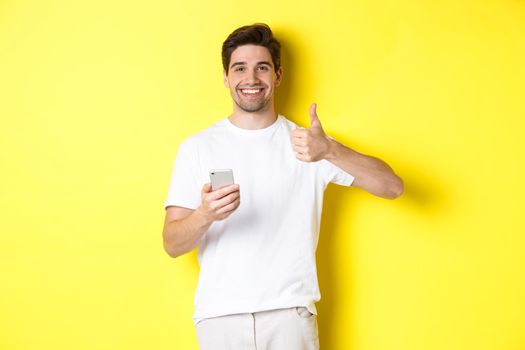 Happy satisfied man holding smartphone, showing thumb up in approval, recommend something online, standing over yellow background.