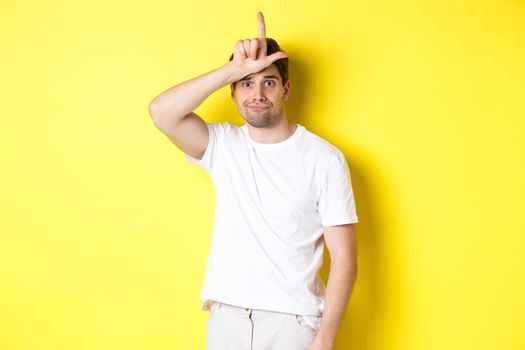 Awkward guy showing loser sign on forehead, looking sad and gloomy, standing in white t-shirt against yellow background.