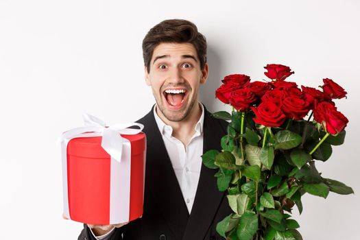 Close-up of handsome bearded man in suit, holding present and bouquet of red roses, smiling at camera, standing against white background.