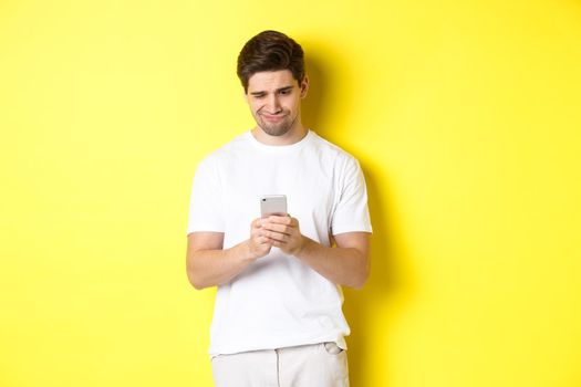 Guy looking displeased at smartphone screen, reading strange message on phone, standing in white t-shirt against yellow background.