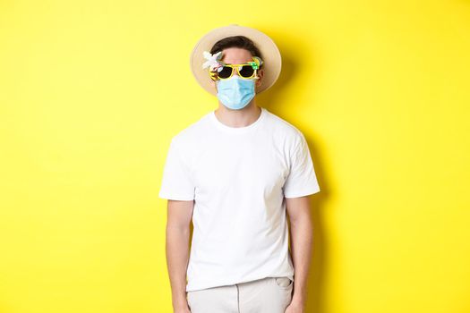 Concept of covid-19, vacation and social distancing. Man tourist wearing medical mask and summer hat with sunglasses, going on trip during pandemic, yellow background.