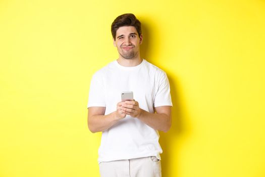 Displeased and reluctant man grimacing, being unamused by message on smartphone, standing over yellow background.
