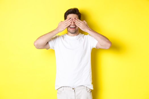 Happy man close eyes and waiting for surprise, smiling amused, standing over yellow background.