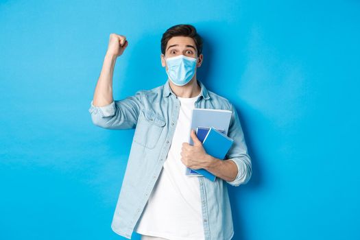 Education, covid-19 and social distancing. Excited male student in medical mask triumphing, raising fist up and holding notebooks, standing over blue background.