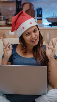 Person talking to family on video call conference to celebrate christmas eve. Woman wearing santa hat while using online conference for remote holiday festivity on laptop in festive room